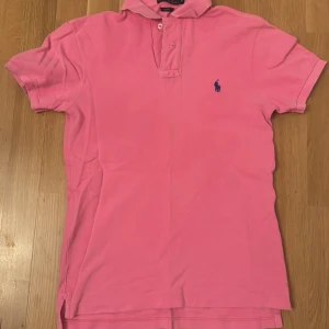 Rosa pikétröja från Ralph Lauren - Säljer en klassisk rosa pikétröja från Ralph Lauren med korta ärmar och blå logga broderad på bröstet. Tröjan har krage och knappar framtill. Perfekt för en avslappnad och stilren look.