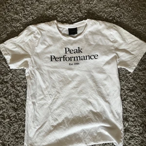 Vit t-shirt från Peak Performance - Säljer en klassisk vit t-shirt från Peak Performance med svart tryck på bröstet. T-shirten har rund halsringning och korta ärmar. Perfekt för en clean och sportig look.