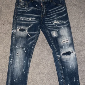Blå jeans från Dsquared2 - Säljer ett par blå jeans från Dsquared2 med slitna detaljer och färgstänk för en edgy look. Jeansen har en klassisk femficksdesign och är tillverkade i Italien. Perfekta för dig som vill ha en unik stil!