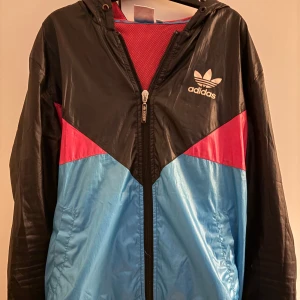 Adidas vindjacka med färgblock - Säljer en snygg vindjacka från Adidas med svart, blå och röd färgblockdesign. Jackan har huva, dragkedja framtill och två fickor. Perfekt för blåsiga dagar och sportig stil. Adidas-logga på bröstet och meshfoder på insidan.