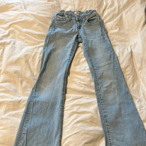 Ljusblå bootcut jeans med fickdetaljer, lite baggy  - Säljer ett par ljusblå jeans med bootcut passform och snygga fickor med knappdetaljer bak. Klassisk femficksmodell och normalhög midja. Perfekta för dig som gillar en avslappnad men ändå trendig stil.