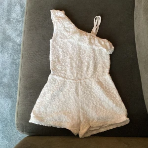 Vit spetsig oneshoulder jumpsuit från Lindex Kids - Superfin vit jumpsuit från Lindex Kids med spetsdetaljer och oneshoulder-design. Perfekt för sommarens varma dagar eller speciella tillfällen. Mjuk insida och elastisk midja för extra komfort.