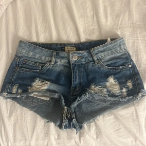Blå slitna jeansshorts från Daysie - Snygga blå jeansshorts från Daysie med slitna detaljer och fransig kant. Klassisk femficksmodell med låg midja och knappgylf. Perfekta för varma dagar och en avslappnad stil.