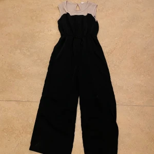 Svart jumpsuit med ribbad överdel - Säljer en svart jumpsuit med breda ben och ärmlös design. Överdelen är ribbad i ljusgrått med rund halsringning och en liten öppning bak. Perfekt för dig som vill ha en stilren och bekväm look.