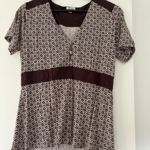 Brunmönstrad blus från Share - Säljer en brun och vit kortklänning/blus från Share med geometriskt mönster. Klänningen har v-ringning, korta ärmar och markerad midja med enfärgade bruna partier. Perfekt för dig som gillar unika mönster och en bekväm passform.