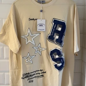 Beige t-shirt från Aelfric Eden - Beige t-shirt från Aelfric Eden med stora broderade stjärnor. Helt ny. Baggy passform i storlek M men passar även L☺️.                                    Hör gärna av dig vid funderingar eller frågor