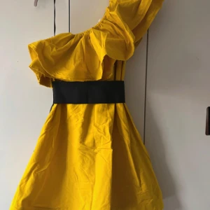 Gul oneshoulder-klänning från Lanvin x H&M - Säljer en statement gul klänning från Lanvin x H&M-kollektionen. Klänningen har en bred svart midjedetalj, volang upptill och en axel. Dragkedja i sidan. Perfekt för dig som vill sticka ut!