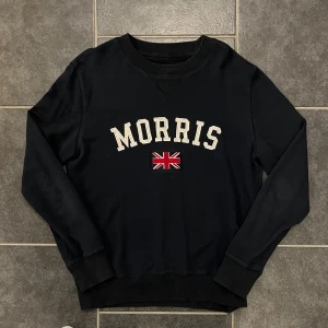 Svart sweatshirt från Morris - Säljer en svart sweatshirt från Morris med broderad logga och brittisk flagga på bröstet. Tröjan har rund halsringning och långa ärmar. Perfekt för en stilren och avslappnad look.