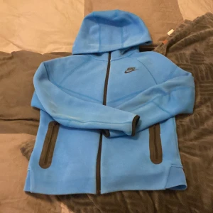 Blå hoodiejacka från Nike - Säljer en blå hoodiejacka från Nike med svarta detaljer och dragkedjor. Jackan har huva, två sidofickor med dragkedja och en extra ficka på ryggen. Perfekt för dig som gillar sportig stil.