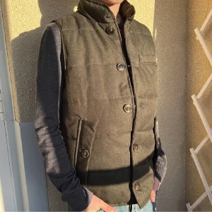 Stilren väst från John Henric - Snygg olivgrön/grå vadderad väst från John Henric med hög krage och knäppning med stora knappar. Västen har två snedställda fickor med lock och knapp samt en bröstficka. Perfekt för lager-på-lager-stil.