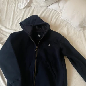 Mörkblå hoodie med dragkedja från Polo Ralph Lauren - Säljer en mörkblå hoodie från Polo Ralph Lauren med dragkedja och klassisk logga på bröstet. Tröjan har huva och långa ärmar, perfekt för en avslappnad stil.