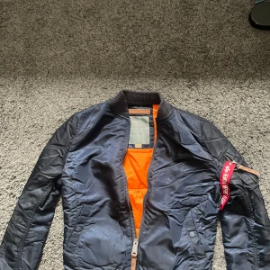 Mörkblå bomberjacka från Alpha Industries - Säljer en klassisk mörkblå bomberjacka från Alpha Industries i storlek small. Jackan har orange foder, ribbade muddar och dragkedja framtill. Detalj med röd tag på ärmen. Perfekt för dig som söker en snygg vårjacka