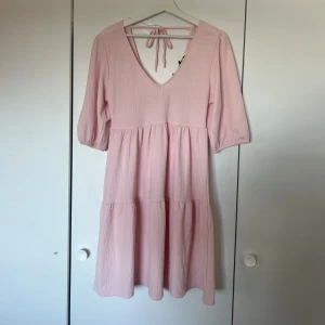 Ljusrosa klänning  - Söt ljusrosa klänning med v-ringning och knyt i ryggen. Klänningen har trekvartsärmar och en luftig, volangig kjol som ger en avslappnad look. Perfekt för varma dagar eller sommarens tillställningar. Den är i väldigt bra skick då den aldrig är använd och prislappen finns fortfarande kvar!🫧✨🩷