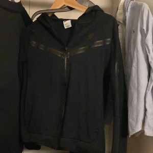 Svart Nike tech med dragkedja - Svart hoodiejacka från Nike med hel dragkedja framtill och svarta detaljer i form av tejpade ränder över bröstet och på ärmen. Jackan har huva och en ficka med dragkedja på ena ärmen. Perfekt för en sportig stil.