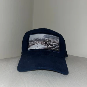 Mörkblå truckerkeps med tryck från Vernando - Säljer en mörkblå truckerkeps från vernando med mesh på sidorna och baksidan. Framtill finns ett svartvitt tryck med motiv och logga. Justerbar storlek baktill med plastspänne. Perfekt för en avslappnad stil.
