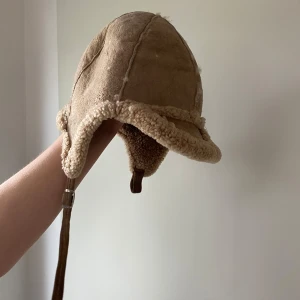 Fårskinnsmössa - Mössa i äkta fårskinn med tryckknapp i spännet. Innermått 56 cm i omkrets. Färg är brun/beige. Köparen står för frakt. 