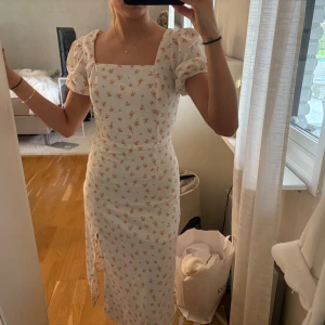 Vit blommig klänning med puffärm och rosett i ryggen - Söt vit klänning med små blommor i rosa och gult. Klänningen har puffärmar, fyrkantig ringning och en fin rosettdetalj i ryggen med öppen rygg. Endast änvänd några få gånger 😍Är i storlek 36 men är ganska liten i storlek så passar 34 också!