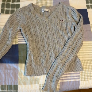 Grå kabelstickad tröja från Hollister - Säljer en grå kabelstickad tröja från Hollister med v-ringning och lång ärm. Tröjan har en liten broderad logga på bröstet och ribbade muddar vid ärmslut och nederkant. En jätte fin tröja!