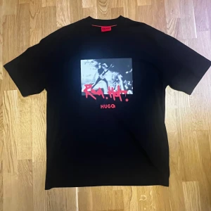 Boss t-shirt - Säljer en svart ”domenade” t-shirt från Hugo Boss med stort grafiskt tryck i vitt och rött framtill. Perfekt för dig som vill sticka ut med en snygg design. 10/10 skick, sparsamt använd, inga skador eller fläckar.