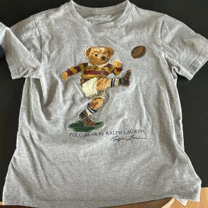 Grå t-shirt med Polo Bear från Ralph Lauren - Grå t-shirt från Ralph Lauren med ikoniska Polo Bear som spelar fotboll tryckt på framsidan. 