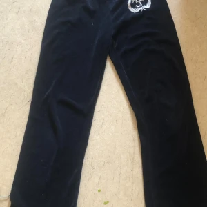 Svarta velourbyxor med gulddetaljer från Juicy Couture - Säljer ett par svarta velourbyxor från Juicy Couture med guldfärgade ränder och knappar i midjan. Byxorna har broderad logga framtill och är perfekta för en avslappnad och trendig look.