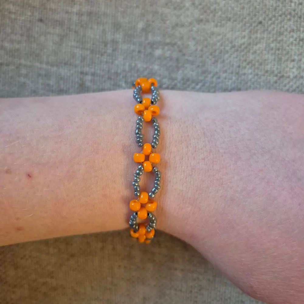 Ge din outfit en färgklick och en unik touch med detta handgjorda pärlarmband, dekorerat med ett charmigt blommönster i lysande orange och skimrande grå pärlor. Den lekfulla och iögonfallande designen passar perfekt för att lyfta vardagslooken eller komplettera sommarens stil.. Asusteet.