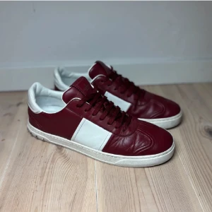 Vinröda och vita sneakers från Valentino - Säljer ett par snygga sneakers från Valentino i vinrött och vitt skinn. Skorna har klassisk snörning, rund tå och vit platt sula. Perfekta för dig som vill ha en stilren och exklusiv look med en sportig touch.