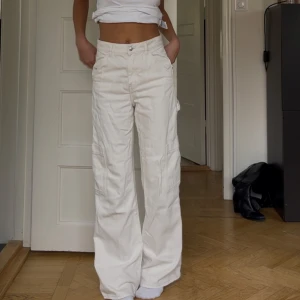 Vita cargopants med raka ben - Säljer ett par vita cargopants med raka, vida ben och flera fickor på sidorna. Byxorna har hög midja och är i bra skick endast med slitningar längst ner på benen pga långa och därför har de slitits mot marken!