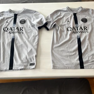 PSG fotbollströjor Neymar Jr och Mbappé - Säljer två grå PSG fotbollströjor med svarta detaljer och tryck. Ena har Neymar Jr 10 på ryggen och den andra Mbappé 7. Båda har Qatar Airways som sponsor och PSG-logga på bröstet. Perfekt för fans av laget!