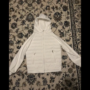 Vit cardigan med huva - Säljer en stilren vit cardigan med huva och dragkedja framtill. Perfekt för våren och enkel att matcha med olika outfits. Är i allmänt mycket bra skick förutom att de har blivit lite färg i innerfickan för jag haft pennor där men syns ej. Lite smutsig också men går att tvätta. Storlek M passar S