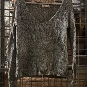 Grå stickad v-ringad tröja från Abercrombie - Mysig grå stickad tröja från Abercrombie med v-ringning och lång ärm. Tröjan har diskret logga broderad på bröstet och ribbade muddar vid ärmslut och nederkant. Perfekt för lager-på-lager-stil. Undom Xl storlek 