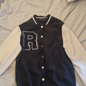 Svart och vit collegejacka med R-tryck - Säljer en klassisk svart och vit collegejacka med stora vita knappar och ett stort R-tryck på bröstet. Jackan har vita ärmar och svart kropp, samt ribbade muddar vid hals och nederkant. Perfekt för en sportig och avslappnad stil.