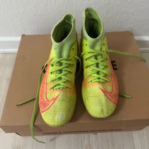 Nike Mercurial fotbollsskor i neongult - Säljer ett par Nike Mercurial fotbollsskor i neongult med röda detaljer och snörning. Skorna har hög krage för extra stöd och är designade för spel på konstgräs. Perfekta för dig som vill sticka ut på planen!