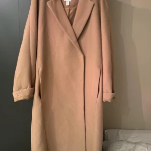Beige kappa från H&M - Säljer en stilren beige kappa från H&M i storlek XS. Jackan har klassisk krage, långa ärmar och knäppning med dolda knappar framtill. Perfekt för dig som gillar en enkel och tidlös look.