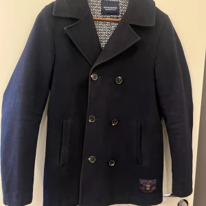 Marinblå skepparkavaj Scotch & Soda - Stilren marinblå skepparkavaj från Scotch & Soda med dubbelknäppning och klassisk krage. Jackan har quiltat innerfoder i gult och två sidofickor. Snygg detalj med broderad patch nertill. (Nypris 2495)!😊