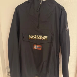 Svart vindjacka från Napapijri - Svart vindjacka från Napapijri med norsk flagga på magen och logotyp på ärmen. Jackan har huva, dragkedja vid halsen och en stor ficka framtill med lock. Perfekt för blåsiga dagar.