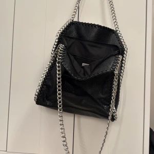Svart axelväska med kedjedetaljer från Tiamo - Cool svart axelväska från Tiamo i syntetmaterial med silverfärgade kedjor som axelremmar och runt kanterna. Väskan har dragkedjefack både inuti och på baksidan, samt en rymlig insida. Perfekt för dig som vill ha en edgy accessoar till din outfit! ⛓️🖤