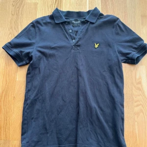 Mörkblå pikétröja från Lyle & Scott och grön från Boomerang - Klassisk svart pikétröja från Lyle & Scott med gul broderad logga på bröstet. Tröjan har krage, tre knappar och korta ärmar. Perfekt för en stilren och avslappnad look.
