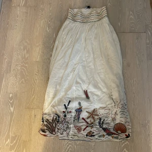 Vit långkjol med broderier från Zara - Vit långkjol från Zara med smock i midjan och färgglada broderier av havsmotiv längst ner. Kjolen har gröna och orangea detaljer i midjan och är tillverkad i ett lätt material. Perfekt för dig som gillar unika detaljer och bohemisk stil. Säljer för att den är för kort för mig