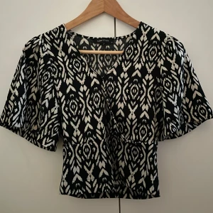 Svartvit mönstrad topp från LCW Vision - Säljer en snygg svartvit mönstrad topp. Toppen har korta ärmar och en v-ringad design som ger en stilren look. Perfekt för att piffa upp garderoben! Har använt några gånger men är i perfekt skick den är lite öppen där framme men jag brukar nåla om man inte vill att den ska vara så öppen 💗