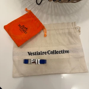 Blått armband från Hermès - Snyggt armband från Hermès i silver med blå detaljer. Armbandet har en elegant design med ett H-format spänne. Perfekt för att ge en touch av lyx till din stil. Vid köp medföljer en hermés dustbag och även en dustbag från vestiaire collective. Självklart äkta och äkthets bevis finns. Vid frågor är det bara att höra av er! En tråkig sak är att piggen till fästet har gått av därav priset