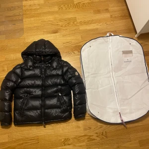 Svart dunjacka från Moncler - Säljer en snygg svart dunjacka från Moncler. Jackan har en pufferdesign med en praktisk huva och dragkedja framtill. Perfekt för kyliga dagar och håller dig varm och stilren. Inkluderar en vit Moncler-förvaringspåse. Knappt använd pga för stor för mig. Köpt begagnad innan men inga håll eller annat förutom små vita fläckar längst ner på högra armen som knappt syns.