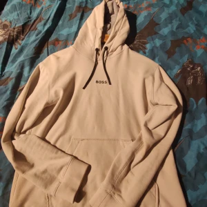 Beige hoodie från BOSS - Säljer en stilren beige hoodie från BOSS med svart logga på bröstet. Den har en klassisk känguruficka och justerbar huva med svarta snören. Perfekt för en avslappnad look. Inga hål eller fläckar osv. Den sitter lite tajt så M/S. Pris kan diskuteras.