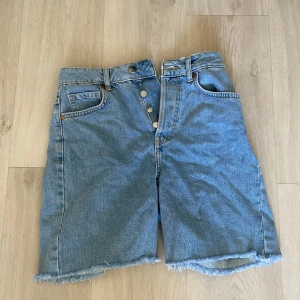 Blå jeansshorts från Bik Bok - Säljer ett par blå jeansshorts från Bik Bok med fransiga kanter och knappar framtill. Perfekta för en avslappnad stil. Tillverkade i 100% bomull för en bekväm passform.