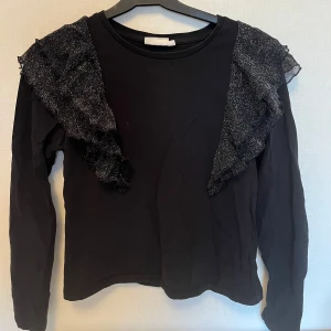 Svart tröja med glittriga volanger från KappAhl - Säljer en svart långärmad tröja från KappAhl med glittriga volanger över axlarna. Perfekt för att lägga till lite glamour i vardagen. Tröjan har en normal passform och är tillverkad i ett mjukt material.