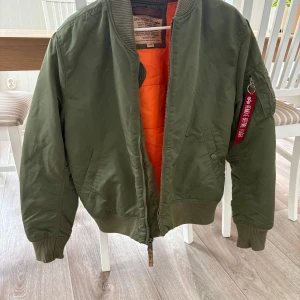 Grön bomberjacka från Alpha Industries - Säljer en grön bomberjacka från Alpha Industries med orange foder. Jackan har en klassisk bomberstil med dragkedja och ribbade muddar. Den har en ikonisk 'Remove Before Flight'-tagg på ärmen. Perfekt för en cool och avslappnad look.