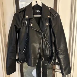Svart skinnjacka från Zara - Snygg svart skinnjacka från Zara med coola dragkedjedetaljer och bälte i midjan. Jackan har en klassisk bikerstil med långa ärmar och knappar på axlarna. Perfekt för att ge din outfit en edgy touch. Storlek M och är markerad med en triangel. 