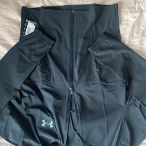 Svart träningsjacka från Under Armour - Säljer en svart träningsjacka från Under Armour med dragkedja och huva. Jackan har långa ärmar och är perfekt för träning. Den är tillverkad i ett syntetmaterial som andas.  Helt ny med prislapp aldrig använd