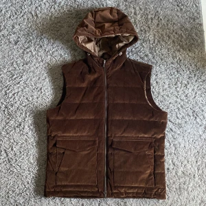 Brun manchester dunväst med huva - Snygg brun Stenstromsväst i manchester med huva och dragkedja. Helt ny! Perfekt för kyligare dagar. Västen har praktiska fickor framtill och en stilren design. Prutning vid snabb affär