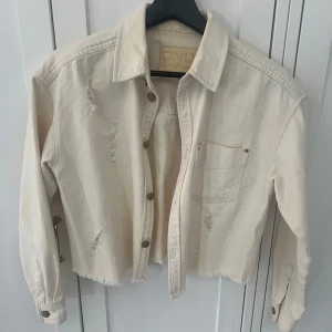 Beige jeansjacka från Only - Säljer en croppad beige jeansjacka från Only med slitna detaljer och fransig nederkant. Jackan har en bröstficka och knappar framtill. Perfekt för en avslappnad stil. 